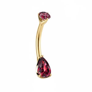 May include: A gold-coloured belly button ring with a curved design. It features a round, red gemstone at the top and a teardrop-shaped, red gemstone at the bottom. The jewellery is set against a white background.