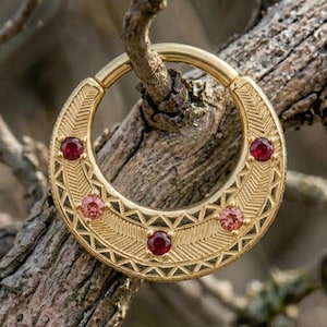 May include: A circular gold hoop earring featuring an intricate engraved chevron pattern and five inset round gemstones. The stones alternate between deep red and lighter pink hues, set symmetrically along the front face of the polished metal band.