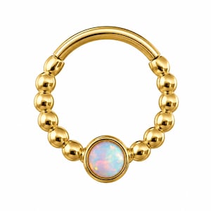 Pode incluir: Um piercing de septo dourado com uma pedra central opalescente redonda. O piercing é adornado com pequenas contas espaçadas uniformemente ao longo de sua superfície curva. A marca "ARGON JEWELS" está impressa na parte inferior.