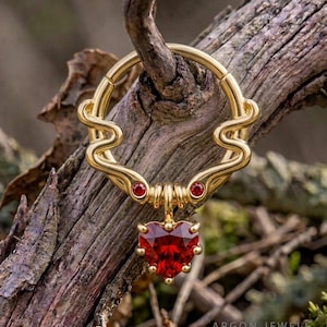 Könnte beinhalten: Ein goldfarbener, kreisförmiger Ring mit einem wellenförmigen Design in der oberen Hälfte, zwei kleinen roten runden Edelsteinen und einem zentralen, herabhängenden herzförmigen roten Edelstein. Der Text Argon Jewels ist unten rechts sichtbar.