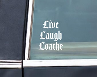 May include: A white vinyl decal applied to the rear window of a black hearse. The decal features the text Live Laugh Loathe in a gothic-style font. A silver cross emblem is also visible on the glass, and a silver ornament is on the side panel.