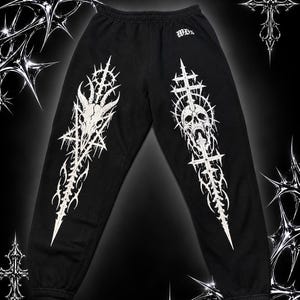 Peut inclure: Pantalon de jogging noir avec des motifs graphiques blancs sur chaque jambe. La jambe gauche présente un crâne de chèvre dans une étoile, tandis que la droite montre un crâne avec une croix. Le texte WD's est imprimé près de la taille.