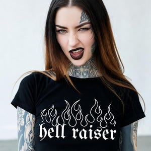 May include: Black crop top with white flame graphic and the words "hell raiser". The model has tattoos on the face, neck, and arms. The model is wearing black lipstick and has dark hair.