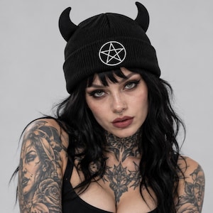 May include: A black ribbed knit beanie featuring two upright horn-shaped protrusions on the crown and a circular patch on the front displaying a white pentagram symbol outlined in black. The hat is constructed from a solid black fabric material.