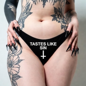 Könnte beinhalten: Schwarze Unterwäsche mit dem weißen Text TASTES LIKE SIN und einem weißen umgedrehten Kreuzsymbol in der Mitte auf der Vorderseite. Das Kleidungsstück besteht aus einem einfarbigen schwarzen Stoff mit schlichtem Schnitt für die Hüfte.