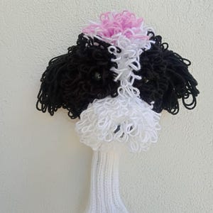 May include: A white and black crocheted golf club head cover shaped like a dog with a pink topknot.