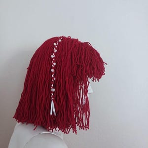Può includere: Parrucca rossa fatta a mano con filato, in stile corto e sfilacciato. La parrucca presenta una treccia di perline bianche e argentate con nastri bianchi. Esposta su una testa di manichino, sfondo bianco.
