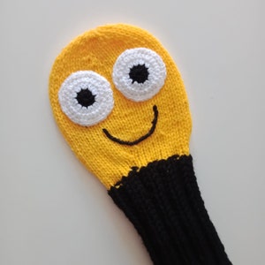 May include: A yellow knitted golf club head cover with a smiling face, black eyes, and white pupils.