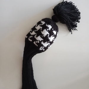 May include: Black and white knitted golf club head cover with a black tassel. The cover has a white pattern of geometric shapes.