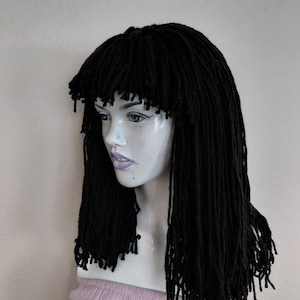 May include: A black wig with long, thick dreadlocks and a blunt fringe. The wig is displayed on a mannequin head with pale skin and a pink top. The dreadlocks are evenly spaced and hang down to the shoulders.