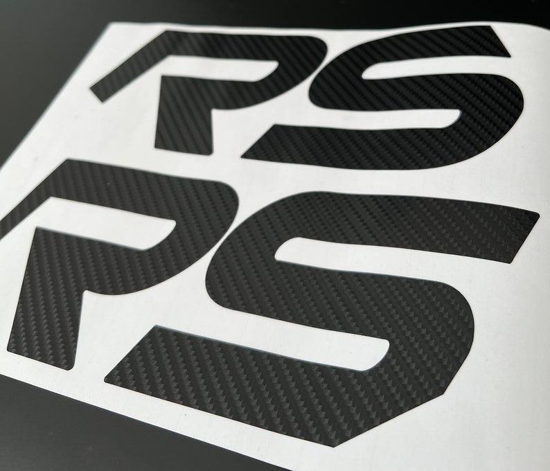 May include: Black carbon fiber vinyl decal with the letters "RS" in a bold, stylized font. The decal is designed to be applied to a car or other vehicle.