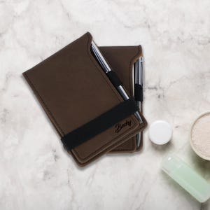 May include: Two brown leather notebook covers with black elastic bands and silver pens. The top cover has the name "Becky" embossed on it.