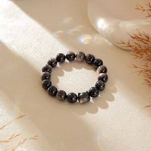 May include: A beaded bracelet composed of smooth, dark grey, reflective stone beads. The bracelet features a single focal bead carved into the shape of a cat head with two small pointed ears, integrated into the circular strand of round beads.
