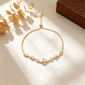 May include: A gold-tone adjustable bracelet featuring a central cross charm encrusted with clear crystals, flanked by two smaller decorative scroll motifs and two round clear crystals on each side, connected by a delicate gold-tone chain.