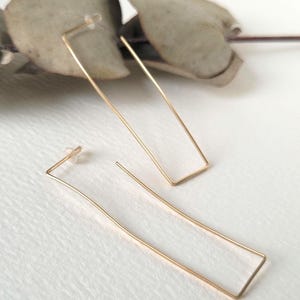 May include: A pair of gold-toned, open rectangle shaped earrings. The earrings are made of thin metal wire and have a slightly curved shape.
