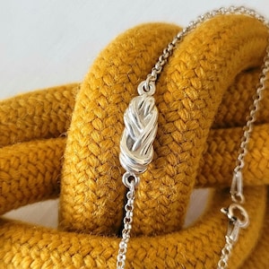 Könnte beinhalten: Eine silberne Halskette mit einem Knotenanhänger, vor einem strukturierten, senfgelben Hintergrund. Die Kette ist filigran, und der Knoten ist aufwendig gestaltet. Der Hintergrund ist ein dickes, gewebtes Seil.