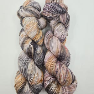 Peut inclure: Deux écheveaux de fil teints à la main dans une palette de couleurs neutres. Le fil présente un mélange de teintes grises, beiges et brunes avec de subtiles touches de rose et de violet. Le fil est bien enroulé, mettant en valeur sa texture.