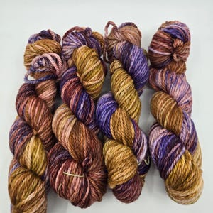 May include: A selection of colourful yarn skeins in shades of brown, purple, and gold. The yarn is arranged in a neat, organised display, ideal for knitting or crocheting projects. The yarn has a textured appearance.