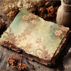 May include: An open, antique-style journal with aged, textured pages. The top page features a floral design in shades of beige, pink, and green. A leather strap and dried flowers are visible, suggesting a vintage aesthetic.