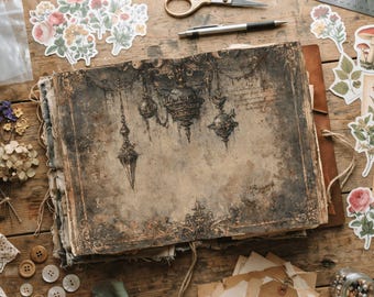 May include: A rectangular journal with a weathered, dark brown cover featuring an intricate illustration of hanging ornate lanterns and decorative borders. The journal is surrounded by floral stickers, a metal ruler, scissors, a pen, and buttons.