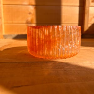 May include: An orange, ribbed glass bowl sits on a wooden surface. The bowl has a cylindrical shape with vertical ridges. The lighting creates shadows, highlighting the texture and colour of the glass. The background is a wooden structure.
