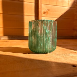 May include: A cylindrical, green glass vase with vertical ribbed detailing. The vase has a marbled appearance with shades of green and sits on a wooden surface. The background is a wooden structure, with sunlight casting shadows.