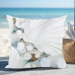 May include: A square throw pillow featuring a printed design of white seashells and smooth, rounded white stones. The pattern displays a collection of fan-shaped shells and circular pebbles in shades of white, soft gray, and muted blue tones.