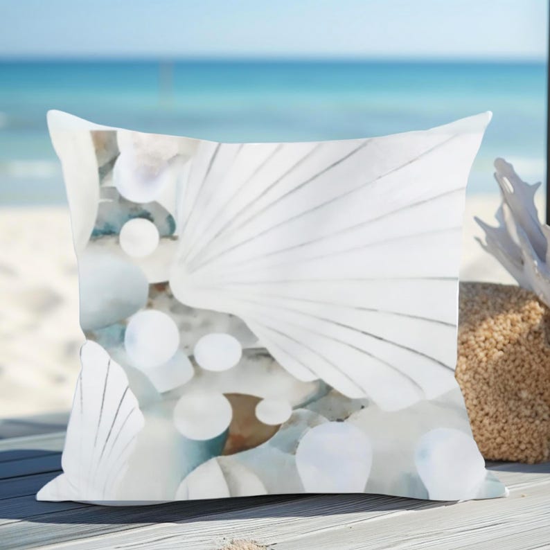 May include: A square throw pillow featuring a printed design of white seashells and smooth, rounded white stones. The pattern displays a collection of fan-shaped shells and circular pebbles in shades of white, soft gray, and muted blue tones.