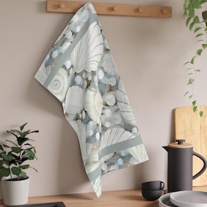 Könnte beinhalten: Ein Küchentuch mit Muschelmuster. Das Tuch zeigt weiße Muscheln und hellblaue Blasen auf hellblauem und braunem Hintergrund. Ein hellgrauer Streifen umrandet das Tuch. Das Tuch hängt an einem Holzregal.