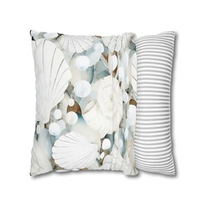 Może przedstawiać: Dwie poduszki dekoracyjne. Jedna kwadratowa poduszka z wzorem muszli w kolorze białym, niebieskim i brązowym. Druga to długa, prostokątna poduszka w szare i białe paski. Poduszki są na białym tle.
