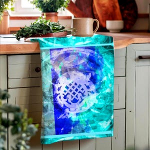 May include: A teal and blue kitchen towel with a white design of a stylized floral pattern. The towel is hanging on a white cabinet door.
