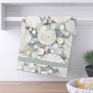 May include: A rectangular kitchen towel featuring a repeating pattern of white seashells and translucent light blue bubbles against a soft background. A solid horizontal gray stripe runs across the lower section of the fabric, which is draped over a white bar.