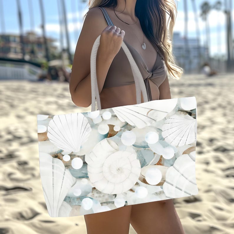 May include: A large tote bag featuring a repeating pattern of white seashells and smooth white pearls against a soft blue and tan background. The bag has two thick white rope handles attached to the top, providing a sturdy grip for carrying items.