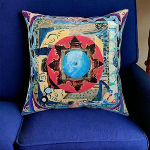 May include: A colorful throw pillow with a geometric design featuring a blue circle surrounded by a red, black, and gold mandala. The pillow has a yellow, blue, and green background with white clouds and other abstract shapes.