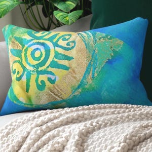 Può includere: Un cuscino rettangolare caratterizzato da uno sfondo vibrante in stile acquerello blu e verde acqua. Al centro è presente un motivo solare stilizzato in giallo brillante e verde con accenti dorati testurizzati, creando un contrasto visivo.