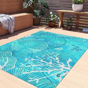 May include: A rectangular outdoor rug with a vibrant turquoise and white seashell design. The rug features a detailed pattern of stylized seashells and coral against a bright turquoise background. The rug is placed on a wooden deck, suggesting outdoor use.