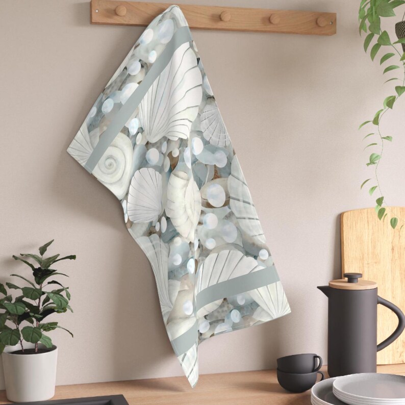 May include: A rectangular kitchen towel featuring a repeating pattern of white seashells and smooth, rounded stones in shades of light blue, gray, and cream. The towel has a soft, muted color palette and is draped over a wooden wall-mounted rack.