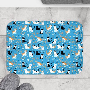 May include: A rectangular bath mat featuring a bright blue background with a repeating pattern of small white daisies and various cats in black, white, and orange colors. The cats are depicted in different sitting and lying positions.