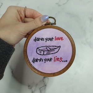 May include: A purple and white embroidery hoop with the text "damn your love" and "damn your lies..." in red and black thread. A small black and white drawing of a boat is in the center of the hoop.