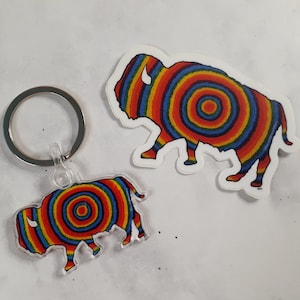 May include: A rainbow-colored bison silhouette keychain with a silver key ring. The bison is made of clear acrylic and has a concentric circle pattern.