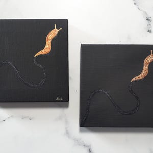 May include: Two square black canvas paintings. Each features a stylized, orange and brown slug with a dark, wavy trail. The slug is positioned in the upper right corner of the canvas. The artist's signature is in the lower right corner.