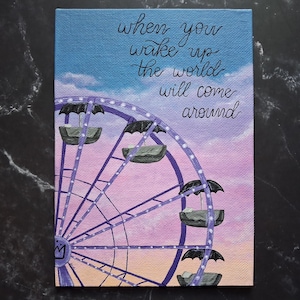 May include: A rectangular canvas painting featuring a purple Ferris wheel with black umbrella-topped passenger cars against a sunset sky. The text "when you wake up the world will come around" is written in black cursive script in the upper right corner.