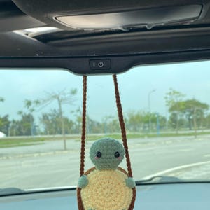 May include: A handmade crochet turtle car charm. The turtle has a green head and limbs, a yellow shell, and a brown border. It hangs from brown crochet strings, suspended from the car's rearview mirror.