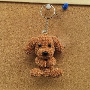 May include: A brown crocheted dog keyring with black eyes and a black nose. The dog is sitting with its paws crossed and is attached to a silver key ring.