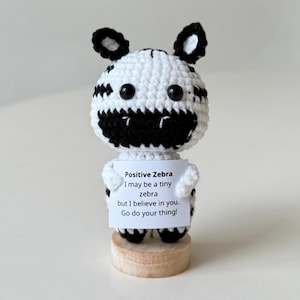 Pode incluir: Uma figura de zebra de crochê com listras pretas e brancas, olhos e orelhas pretas. Segura uma placa que diz "Positive Zebra, I may be a tiny zebra but I believe in you. Go do your thing!" A zebra está em uma base de madeira.
