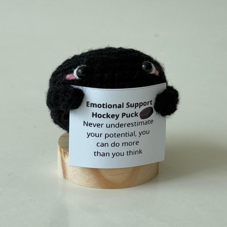 May include: Black felt hockey puck with a cute face holding a sign that says "Emotional Support Hockey Puck Never underestimate your potential, you can do more than you think".