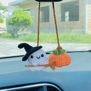 May include: Crocheted Halloween car charm featuring a white ghost wearing a black witch hat and an orange pumpkin. The ghost is holding a broom. The charm is suspended by brown yarn.
