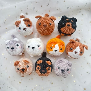 May include: A collection of ten handmade crochet dog toys in various colors and patterns. Each toy is round with a small tail and features a different breed design, including brown, white, black, and gray. The toys are arranged on a white surface with gold star accents.
