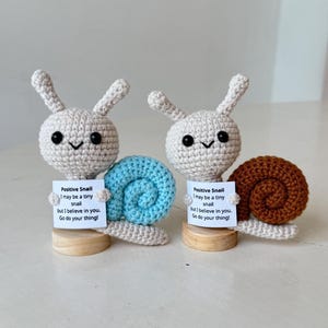 May include: Two crocheted snail figurines. One has a light blue shell, the other a brown shell. Each has a white head with black eyes and a small sign that reads "Positive Snail, I may be a tiny snail but I believe in you. Go do your thing!"