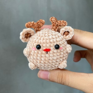 May include: A small, plush, beige reindeer toy with brown antlers and ears. The reindeer has black eyes, a red nose, and pink cheeks. The toy is round and has a soft, textured appearance.